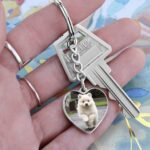 Personalized heart-shaped pet photo keychain in surgical steel, displayed in a hand. Features a custom pet photo engraving on the front with optional custom text engraving on the back