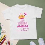 White birthday girl's shirt laid flat, featuring a gold crown graphic above a personalized name in elegant lettering