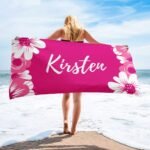Custom towel with name in medium red violet, featuring floral design — perfect beach gift for her, laid out near the shore