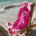 Floral custom beach towel with personalized name in medium red violet color, displayed on a chair at the beach with ocean in background