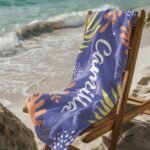 Personalized tropical towel with name for her draped over a wooden chair near the beach – features vibrant tropical leaf design and custom name, ideal for summer vacations or beach getaways