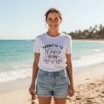 Woman wearing a white Promoted to Mama Est custom year shirt standing on the beach, personalized new mom t-shirt for first time mothers